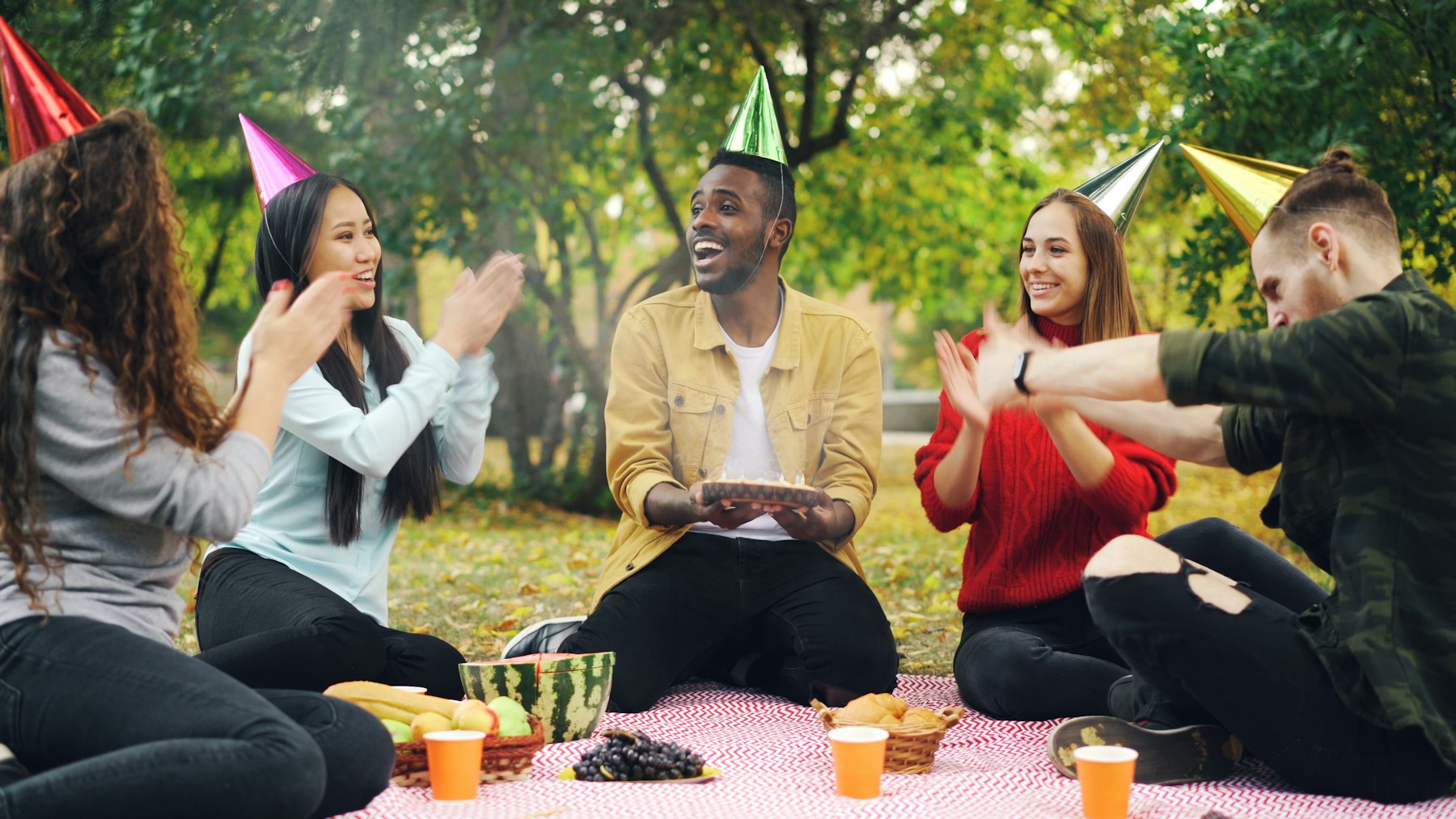 Friends in party hats laughing around an outdoor picnic spread